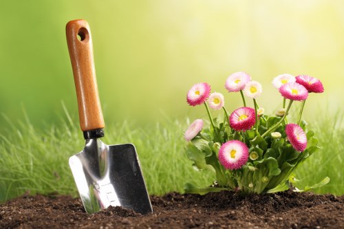 A small garden scene showing a patch of freshly turned dark brown soil in the foreground, with a metal gardening trowel featuring a wooden handle partially inserted into the earth on the left side. To the right, there is a vibrant cluster of pink and white flowering plants with rounded petals, positioned in a lush green garden bed. The background displays a softly blurred grassy lawn with a gradient of green tones, suggesting a bright, sunny day possibly in a suburban garden setting in Barnet. The overall scene highlights careful garden planting and maintenance, consistent with professional gardening services in the Barnet area, and showcases natural landscaping elements suitable for a typical outdoor residential space.