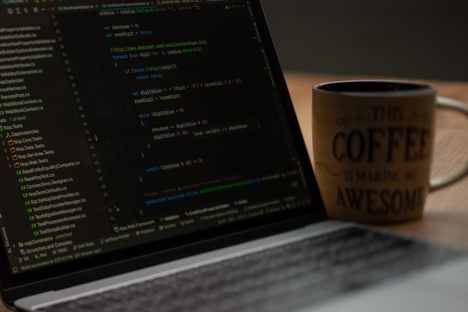 A close-up view of a laptop displaying lines of colorful code on a dark background, resting on a wooden desk. Next to the laptop is a ceramic mug with a beige background and black text that reads 'Thi