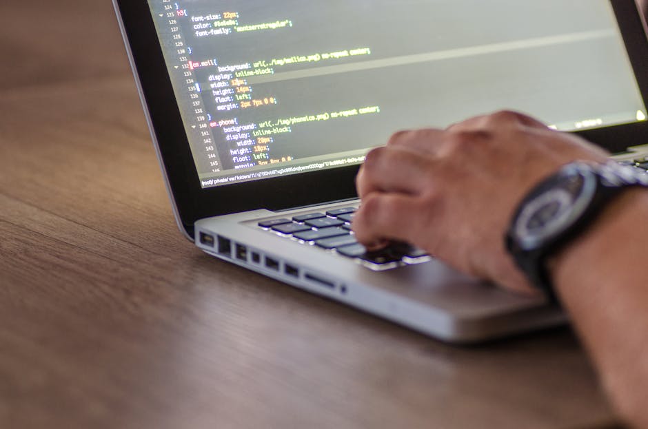 A close-up view of a laptop displaying lines of colorful code on a dark background, resting on a wooden desk. Next to the laptop is a ceramic mug with a beige background and black text that reads 'Thi